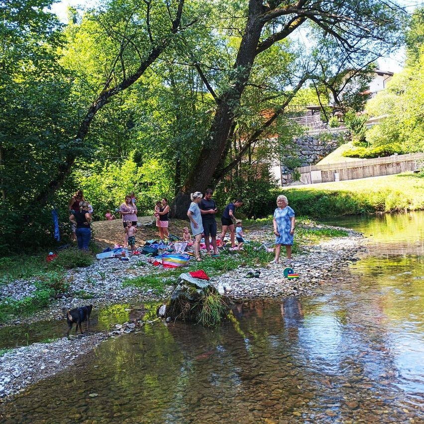 Eine Gruppe von Menschen versammelt sich an einem Fluss, einige stehen und andere sitzen auf dem Grasufer. Ein Hund ist im Wasser, und verschiedene Gegenstände sind verstreut, einschließlich einer Decke und eines Spielzeugs.