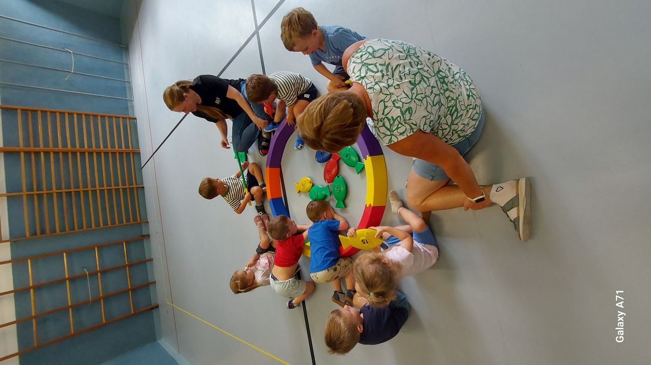 Eine Gruppe von Kindern und zwei Erwachsenen spielt mit einem bunten Fischspielzeug auf einem weißen Boden. Die Kinder sitzen im Kreis um das Spielzeug, während sich die Erwachsenen herunterbeugen, um ihnen zu helfen.