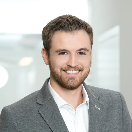 Ein Mann in einem grauen Blazer und weißem Hemd steht lächelnd in einem gut beleuchteten Büro.