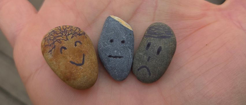 Drei kleine Steine mit gezeichneten Gesichtern, einer lächelnd, einer neutral und einer missmutig, liegen in der Hand einer Person. Die Steine variieren in Farbe und Größe.