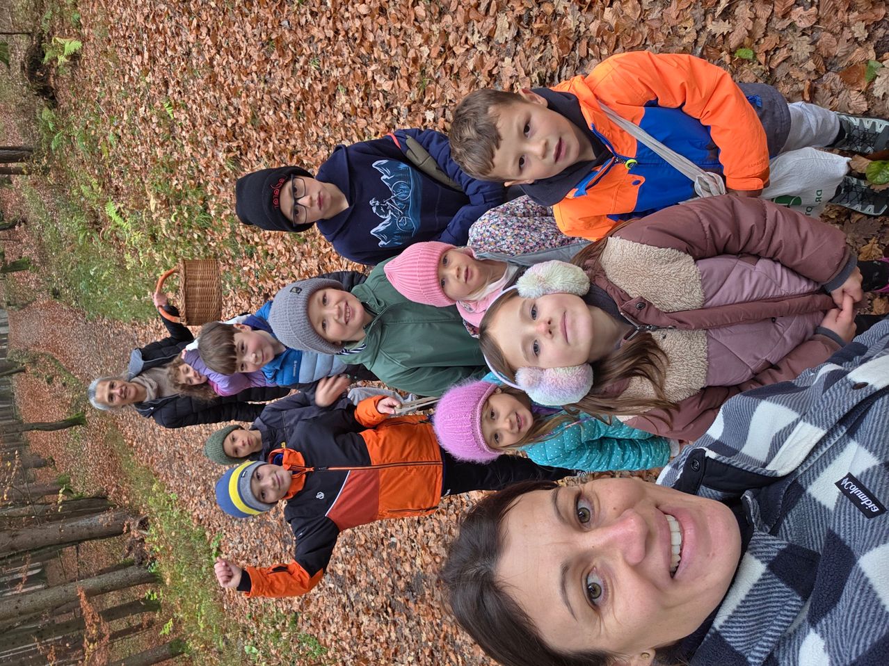 Eine Gruppe von Kindern und einer Frau liegt auf einem mit Blättern bedeckten Waldboden. Sie alle lächeln, wobei ein Kind einen Korb hält.