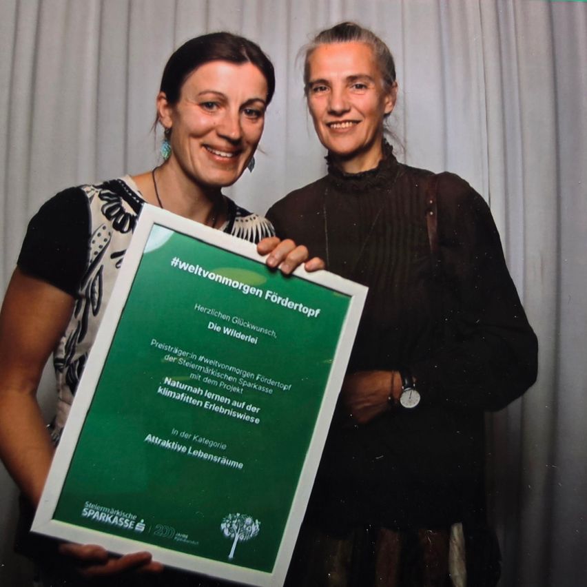 Zwei Frauen stehen lächelnd da, eine hält ein grünes Zertifikat mit Text über klimafreundliches Leben. Sie posieren für ein Foto vor einem weißen Vorhang.