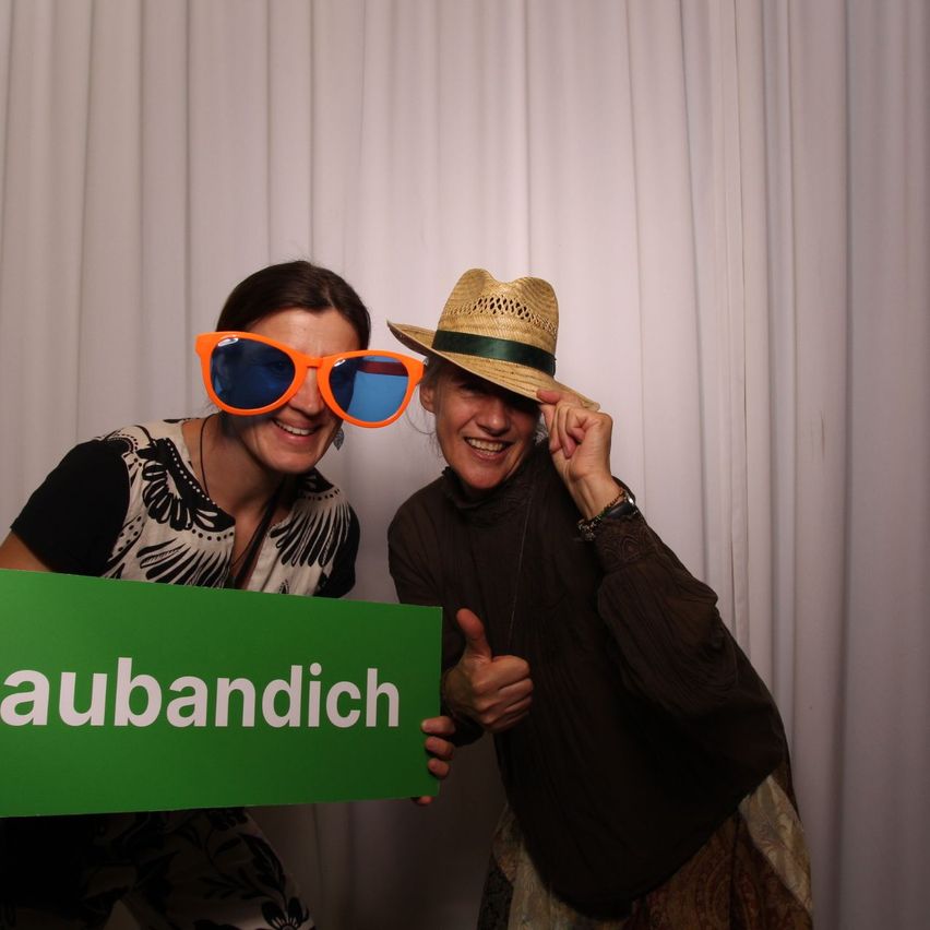 Zwei Frauen lächeln und posieren für ein Foto. Eine hält ein grünes Schild mit weißem Text, der 'aubandich' lautet. Beide tragen Sonnenbrillen und Hüte. Der Hintergrund ist ein weißer Vorhang.