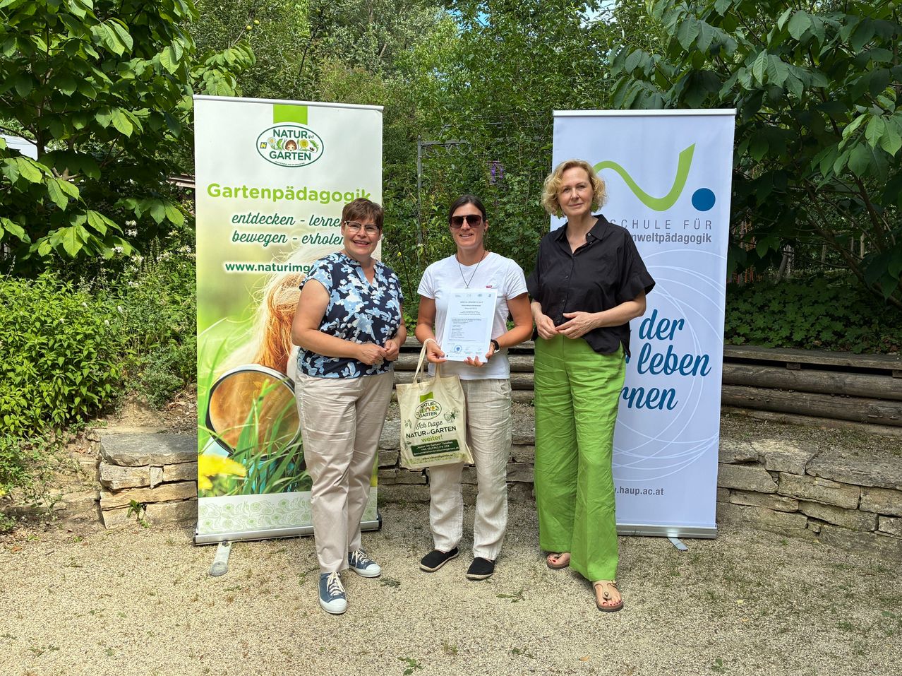 Three women stand in front of banners, one holding a certificate and a bag. They are smiling and appear to be at an outdoor event.