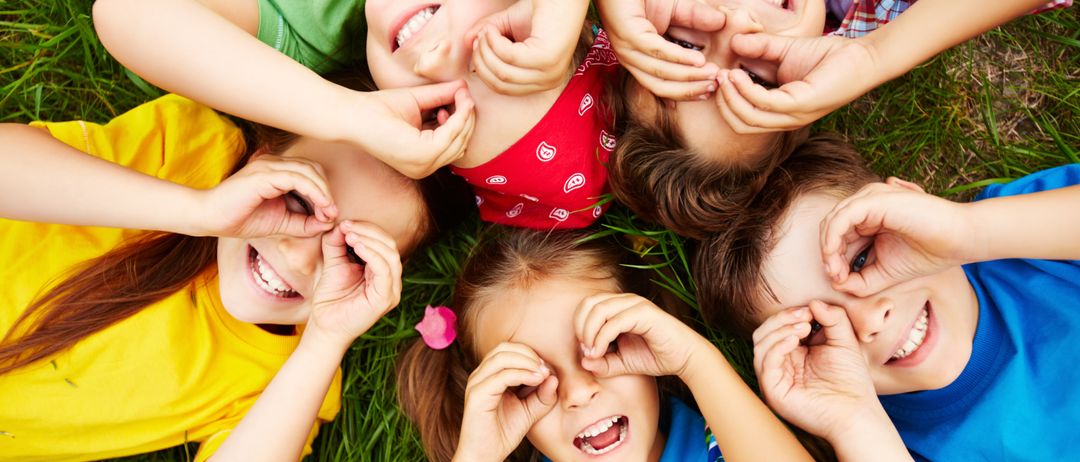 Eine Gruppe von Kindern liegt auf dem Gras, lächelt und schaut durch ihre Finger. Sie tragen alle verschiedenfarbige T-Shirts.