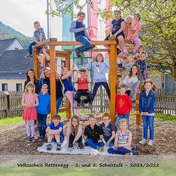Schüler der Volksschule Rettenbach posieren für ein Gruppenfoto auf einem Spielplatz mit Holzkonstruktionen, umgeben von Bäumen und Häusern. Das Foto wurde im Schuljahr 2021/2022 aufgenommen.