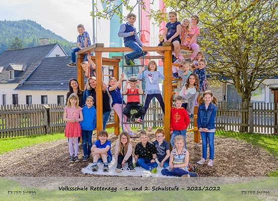 Schüler der Volksschule Rettenbach posieren für ein Gruppenfoto auf einem Spielplatz mit Holzkonstruktionen, umgeben von Bäumen und Häusern. Das Foto wurde im Schuljahr 2021/2022 aufgenommen.
