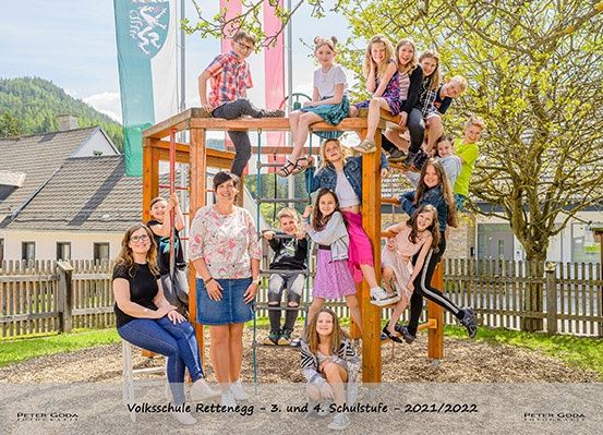 Eine Gruppe von Kindern und zwei erwachsenen Frauen posiert für ein Foto auf einem Spielplatz, umgeben von Holzzäunen, einem Haus und Bäumen. Das Foto wurde 2021/2022 aufgenommen.