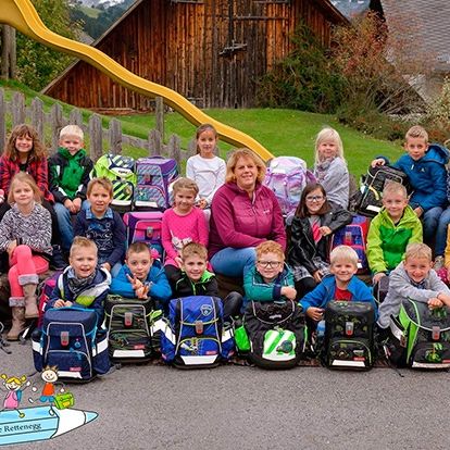 Eine Gruppe von Kindern und einer Frau steht draußen mit ihren Rucksäcken. Die Kinder lächeln und einige schauen direkt in die Kamera. Die Frau steht vor ihnen. Hinter der Gruppe befindet sich ein Haus mit einem Holzzaun, einer Rutsche und Pflanzen.