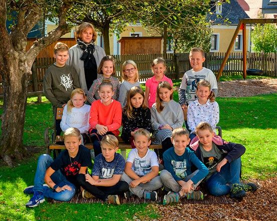 Eine Gruppe von Kindern und ihrer Lehrerin posiert für ein Foto in einem Park mit einem Baum, einer Bank und einem Spielplatz im Hintergrund.