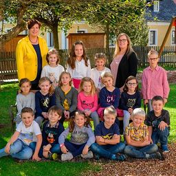 Eine Gruppe von Kindern und zwei Frauen sitzt auf einer Bank im Park und lächelt für ein Foto. Die Frau auf der linken Seite trägt eine gelbe Jacke und die Frau auf der rechten Seite trägt eine Brille. Die Kinder tragen Freizeitkleidung, und einige tragen auch eine Brille.