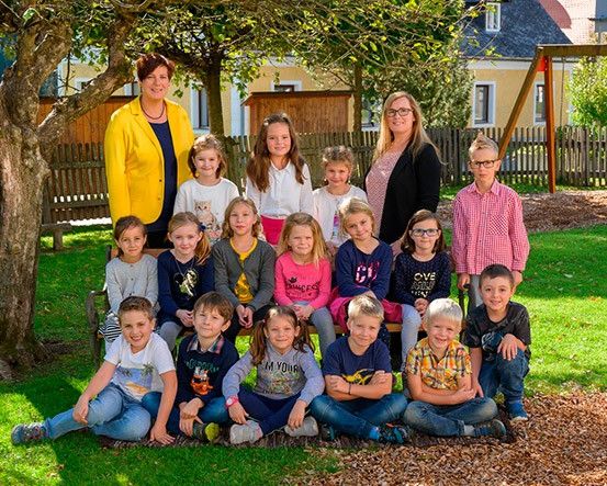 Eine Gruppe von Kindern und zwei Frauen sitzt auf einer Bank im Park und lächelt für ein Foto. Die Frau auf der linken Seite trägt eine gelbe Jacke und die Frau auf der rechten Seite trägt eine Brille. Die Kinder tragen Freizeitkleidung, und einige tragen auch eine Brille.
