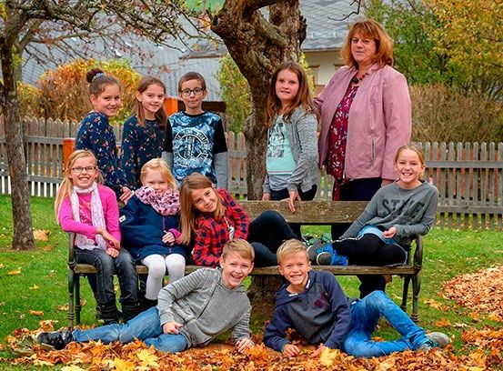 Eine Gruppe von Kindern und einer erwachsenen Frau posiert für ein Foto auf einer Bank im Park. Einige der Kinder liegen auf dem mit Blättern bedeckten Boden. Dahinter befindet sich ein Holzzaun und ein Baum mit einigen Blättern.