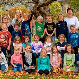 Eine Gruppe von Kindern und ihren Lehrern posiert für ein Foto auf einem Rasen. Die Kinder sind in verschiedenen Farben und Stilen gekleidet. Im Hintergrund befinden sich ein Baum, ein weißer Zaun und ein Haus.