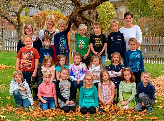 Eine Gruppe von Kindern und ihren Lehrern posiert für ein Foto auf einem Rasen. Die Kinder sind in verschiedenen Farben und Stilen gekleidet. Im Hintergrund befinden sich ein Baum, ein weißer Zaun und ein Haus.