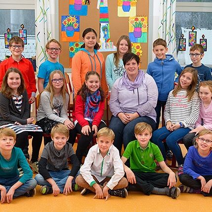 Eine Gruppe von Kindern und einer erwachsenen Frau sitzen zusammen auf einer Bank in einem Klassenzimmer. Das Klassenzimmer hat weiße Wände mit Bilderrahmen und Zeichnungen, und es gibt Regale mit Büchern und Spielzeug.