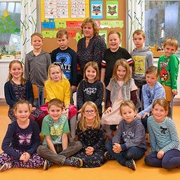 Eine Gruppe von Kindern und ihrer Lehrerin posiert für ein Foto in einem Klassenzimmer mit einem farbenfrohen Bulletin Board und Fenstern.