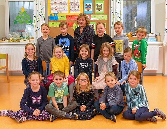 Eine Gruppe von Kindern und ihrer Lehrerin posiert für ein Foto in einem Klassenzimmer mit einem farbenfrohen Bulletin Board und Fenstern.