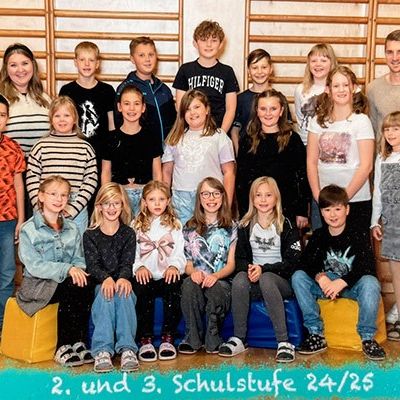 Eine Gruppe von Schülern posiert für ein Foto vor einer Holzwand mit dem Text '2. und 3. Schulstufe 24/25'. Sie sitzen auf gelben Blöcken.