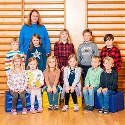Eine Gruppe von Kindern und einer Frau posiert für ein Foto in einem Raum mit Holzwänden und einem Holzboden. Die Kinder sitzen auf blauen Würfeln und die Frau steht hinter ihnen.
