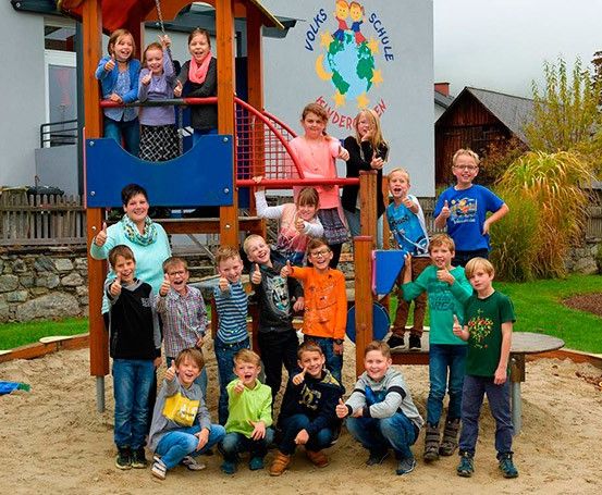 Kinder und Erwachsene vor einem blauen und roten Klettergerüst. Sie lächeln und zeigen Daumen hoch. Dahinter steht ein Gebäude mit dem Schild 'Volksschule'.