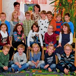 Eine Gruppe von Kindern in einem Klassenzimmer mit einer erwachsenen Lehrerin, die hinter ihnen steht. Die Kinder sitzen und knien auf einem Teppich mit einer Straßengestaltung.