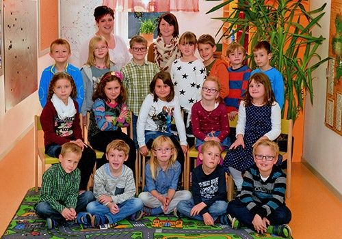Eine Gruppe von Kindern in einem Klassenzimmer mit einer erwachsenen Lehrerin, die hinter ihnen steht. Die Kinder sitzen und knien auf einem Teppich mit einer Straßengestaltung.