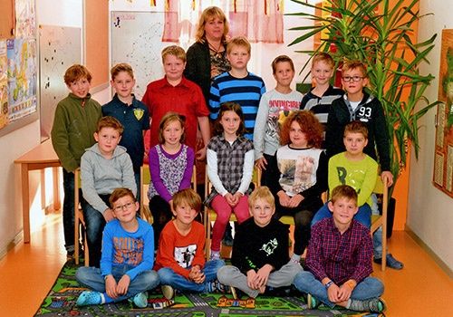 Eine Gruppe von Kindern und einer erwachsenen Frau posiert für ein Foto in einem Klassenzimmer. Die Kinder sitzen und stehen auf einem Teppich, während die Frau hinter ihnen steht.