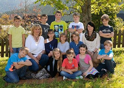 Eine Gruppe von Kindern und zwei Frauen posiert für ein Foto in einem Park mit einem Holzzaun und Bäumen im Hintergrund.
