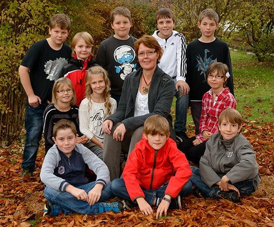 Gruppenfoto einer Frau und sieben Kindern, die auf Herbstblättern sitzen. Die Frau trägt eine Brille und eine Halskette. Die Kinder tragen Freizeitkleidung.