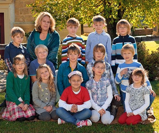 Eine Gruppe von Kindern und einer Frau sitzen auf dem Rasen vor einem Gebäude.