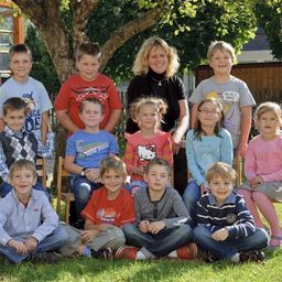 Eine Gruppe von Kindern und einer erwachsenen Frau posiert für ein Foto im Park, mit einem Baum und einem Holzzaun im Hintergrund.
