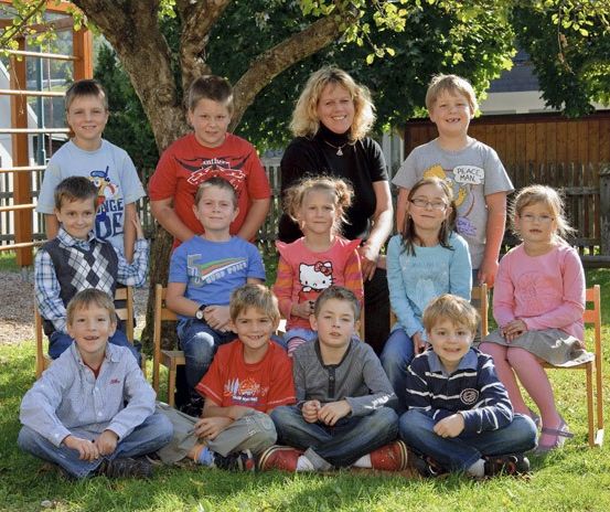 Eine Gruppe von Kindern und einer erwachsenen Frau posiert für ein Foto im Park, mit einem Baum und einem Holzzaun im Hintergrund.