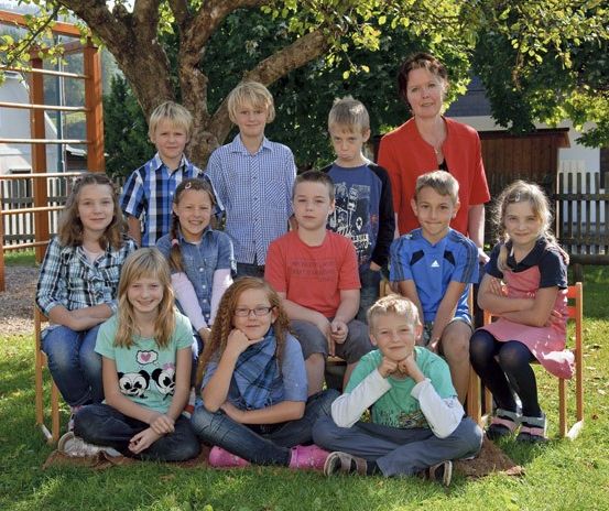 Eine Gruppe von Kindern und einer erwachsenen Frau sitzt auf dem Gras und posiert für ein Foto. Dahinter steht ein Baum, ein Holzzaun und ein Gebäude.