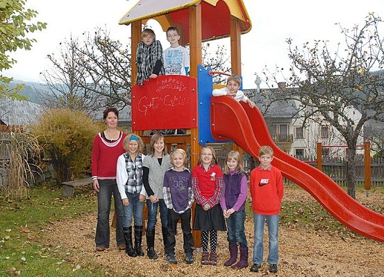Eine Gruppe von Kindern und einer erwachsenen Frau posiert vor einem Spielplatz mit einem roten Rutschen und einem Baum.
