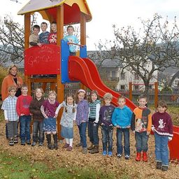 Eine Gruppe von Kindern und zwei Erwachsenen posiert vor einer bunten Spielstruktur mit einer roten Rutsche.