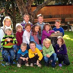 Eine Gruppe von Kindern und einer Frau posiert für ein Foto im Garten mit einem Baum, einem Holzzaun und Bänken im Hintergrund.