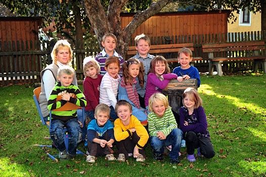 Eine Gruppe von Kindern und einer Frau posiert für ein Foto im Garten mit einem Baum, einem Holzzaun und Bänken im Hintergrund.