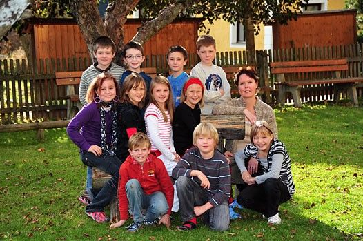 Eine Gruppe von Kindern und einer Frau posiert für ein Foto auf einem Rasen, mit einem hölzernen Baumstumpf, Bänken und einem Zaun im Hintergrund.