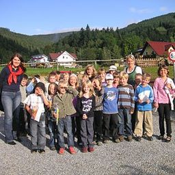 Eine Gruppe von Kindern und ihren Lehrern posiert für ein Foto auf einem Schotterweg, mit einem Spielplatz und Häusern im Hintergrund.
