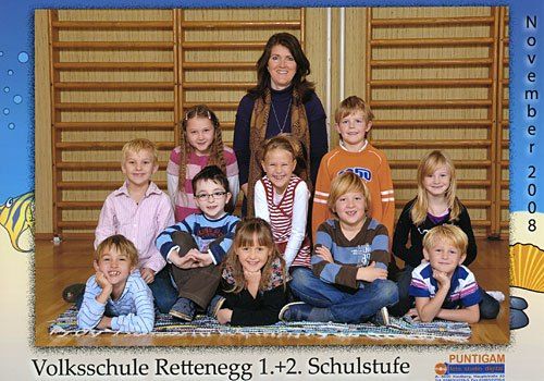 Ein Gruppenfoto der Schüler und ihrer Lehrerin von der Volksschule Rettenegge, aufgenommen im November 2008. Sie sitzen auf einem Teppich und lächeln für die Kamera.