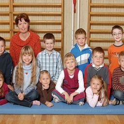 Ein Gruppenfoto von jungen Schülern in einer Turnhalle, die auf einer blauen Matte sitzen, während eine Lehrerin hinter ihnen steht.