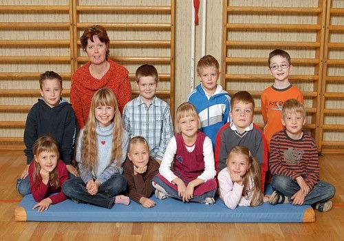 Ein Gruppenfoto von jungen Schülern in einer Turnhalle, die auf einer blauen Matte sitzen, während eine Lehrerin hinter ihnen steht.