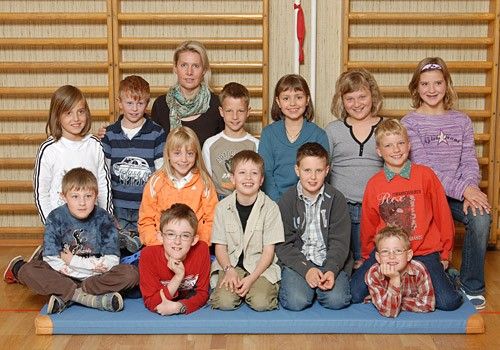Eine Gruppe von Kindern und einer Frau posiert für ein Foto, sitzend auf einer blauen Matte auf einem Holzboden. Die Frau steht hinter ihnen.