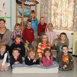 Eine Gruppe von Kindern und einer erwachsenen Frau posiert für ein Foto in einem Klassenzimmer. Sie sitzen und stehen um eine große weiße Matratze herum. Der Raum hat farbenfrohe Gemälde und Dekorationen an den Wänden.