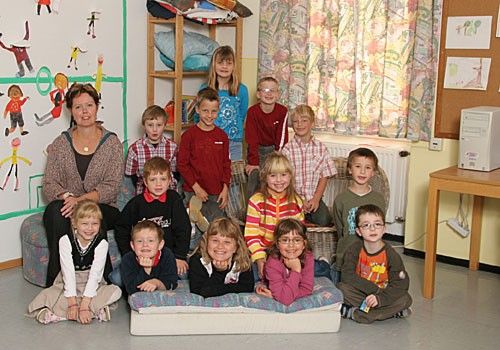 Eine Gruppe von Kindern und einer erwachsenen Frau posiert für ein Foto in einem Klassenzimmer. Sie sitzen und stehen um eine große weiße Matratze herum. Der Raum hat farbenfrohe Gemälde und Dekorationen an den Wänden.