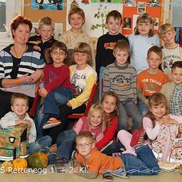Eine Gruppenaufnahme einer Kindergartenklasse mit ihrer Lehrerin im Jahr 2005, sitzend und stehend in Reihen, in Freizeitkleidung, mit einem Halloween-Kürbis und Schulranzen im Hintergrund.