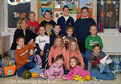 Eine Gruppe von Kindern und zwei Lehrern in einem Klassenzimmer, lächeln für ein Foto. Einige tragen Turnschuhe, Brillen und Armbänder. Ein Kürbis und ein Rucksack liegen auf dem Boden.