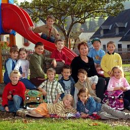 Eine Gruppe von Kindern und Erwachsenen posiert für ein Foto im Spielplatz, mit einer Rutsche und Häusern im Hintergrund.