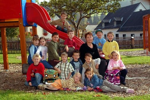 Eine Gruppe von Kindern und Erwachsenen posiert für ein Foto im Spielplatz, mit einer Rutsche und Häusern im Hintergrund.